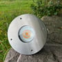 Light By Juul Gold nedgravningsspot big ligger på en sten ved græsset, set forfra med rund stålfront og LED-linse.