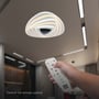 En hånd holder fjernbetjeningen til V-TAC LED loftlampen, mens lampen er monteret i et moderne rum, illustrerende fjernstyringsfunktionen.