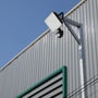 V-TAC LED projektør med PIR sensor monteret på metalstolpe ved en bygning under blå himmel.