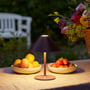 Mørkerød Halo Design Hygge bordlampe oplyser et havebord med frugtskåle, skaber hyggelig stemning.