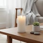 LEDlife hvidt bloklys på 15 cm i ægte stearin, med realistisk flammeeffekt, placeret på et sofabord med planter og sten.