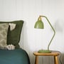 Superliving Urban bordlampe i mat olivengrøn med messingdetaljer står på et lille træbord ved siden af en seng.