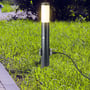 V-TAC pullertlampe monteret i en græsplæne om aftenen, lyser med varmt skær og er tilsluttet strøm via en ledning.
