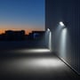 To V-TAC LED-projektører lyser op langs en lagerbygning og ud over en udendørs plads ved skumring.