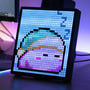 32x32 pixel Bluetooth LED display viser et sovende pixelkunst ansigt med lilla og blå nuancer, placeret på et skrivebord med en computerskærm i baggrunden.