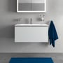 Duravit L-Cube vaskeskab i hvid mat med én skuffe monteret på en grå væg, med et blåt håndklæde hængende til højre.