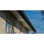 Plastmo sort tagrende monteret på husfacade med lyse mursten og hvide vinduer mod blå himmel.