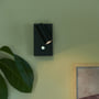 Nordlux Omari væglampe i mat sort metal med justerbart lys på en lysegrøn væg, ved siden af en stor grøn plante og et maleri.