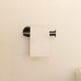 Cosmic toiletpapirholder uden låg i sort mat finish monteret på cremefarvet, tekstureret væg med toiletpapir påsat.