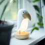 Star Trading Tukanfugl LED børnelampe i hvid, med en træfarvet næbdetalje, placeret på en vindueskarm med planter i baggrunden.