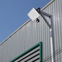 V-TAC LED projektør monteret udendørs på en bygningsfacade med lys grå metalbeklædning og blå himmel.