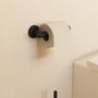 Cosmic Architect S+ toiletpapirholder i mat sort, monteret på en lys væg med en rulle toiletpapir påsat.