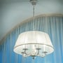 Hvid lampe med stofskærm og dekorative metalarme, der belyser et rum med blå gardiner.