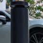 Light By Juul Platin-pullert set tæt på ved en parkeret bil, med rund lampehat og mørk cylindrisk krop.