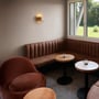 SLV Pantilo Rope væglampe i messingfarvet glas og gyldne metalaccenter, monteret på en væg i et loungeområde med brune sofaer og runde træborde.