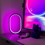 RGBIC ellipseformet lampe, 25 x 15 cm, oplyst med lilla lys på et skrivebord ved siden af en computerskærm.