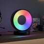Cirkel RGBIC lampe i brug med en regnbuefarvet glød på en hylde ved siden af gamingudstyr og figurer.