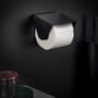 Cosmic toiletpapirholder med låg i børstet sort finish, vist på en mørk væg med en toiletpapirrulle indsat.
