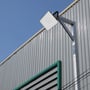 V-TAC 50W LED projektør monteret på stolpe udenfor en industribygning med ribbet facade og blå himmel.