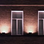 V-TAC LED projektør i brug om natten, oplyser en murstensfacade med to vinduer i varmt, gyldent lys.