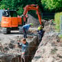 Anlægsstruktørlærlinge i gang med udgravning, assisteret af en orange Hitachi gravemaskine under arbejde på en byggeplads.