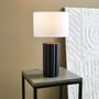 Markslöjd Column bordlampe i sort med en hvid lampeskærm, placeret på et lille bord ved siden af en vægdekoration.