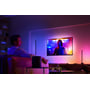 En person ser fjernsyn i et mørkt rum, oplyst af den dynamiske og farverige lyseffekt fra en Philips Hue Lightstrip Gradient.