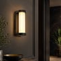 V-TAC LED-væglampe med varmt lys monteret på væg ved en mørk indgang med planter omkring.