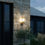 Nordlux Linton udendørs væglampe monteret på en stenmur ved mørkets frembrud, med oplyst klart glas og galvaniseret stål.