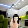 En hånd holder V-TAC touch-fjernbetjeningen til at justere LED lys i et moderne køkkenmiljø med synlige armaturer.