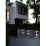 Flere Nordlux Grip Smart hvide LED-væglamper lyser langs en lav betonmur foran et moderne hus og have.