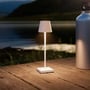 Opladelig hvid LED mini bordlampe på en træterrasse ved vandet om aftenen, med varmt lys.