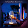 En person tager Philips Hue Go bordlampen med udendørs, hvor den lyser varmt hvidt, mens folk sidder ved et bord.