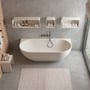 Ideavit Solidnova fritstående badekar i mat hvid solid surface, måler 180x80 cm, placeret mod en betonvæg med hylder og håndklæder.