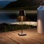 Sort LED mini bordlampe tændt på et træbord ved vandkanten om aftenen, med en stor vandflaske til højre.
