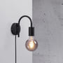 Nordlux Paco retro væglampe i sort metal med E27 fatning og synlig lyskilde, monteret på væg ved siden af en rå betonvæg.