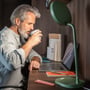 Mand sidder ved skrivebordet med grøn Lumie Task lampe, mens han drikker kaffe og arbejder på laptop.