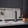 Philips sodamaker i stål med tilhørende flaske placeret på en køkkenbordplade med frugt og glas.