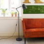 Superliving Dynamo gulvlampe i mat sort metal, vist ved et vindue med planter og en rustfarvet sofa i stuen.
