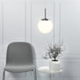 Nordlux Cafe pendel med opal hvid glaskuppel og metalophæng, vist over et lille hvidt bord med en potteplante ved siden af en grå stol.