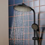Hansgrohe Activera S Showerpipe i sort, med vand der løber fra hovedbruseren og håndbruseren, monteret på en væg med mørkeblå fliser.