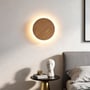 LEDlife væglampe i lyst træ lyser over et natbord ved siden af en seng og fremhæver sin runde form.