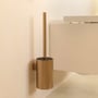 Cosmic Wall toiletbørsteholder i børstet guld, monteret på en cremefarvet væg ved siden af et toilet.