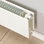 Detalje af hvid Hudevad LK konvektor H210, vist ved siden af en væg med trægulv.
