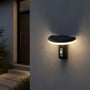 V-TAC LED-solcellehavelampe med sensor lyser på en husmur ved en indgang, vist på afstand i mørke.