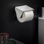 Cosmic toiletpapirholder i mat hvid med låg, monteret på sort væg med en rulle toiletpapir. Synlig del af toiletbørste i baggrunden.