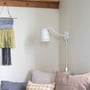 Superliving Dynamo 345 væglampe i mat hvid metal med et fleksibelt drejeled monteret på en lys væg ved siden af en sofa og et vævet vægophæng.