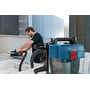 Bosch GAS 18V-10L Professional-støvsuger er koblet til slangen, mens en håndværker arbejder med elværktøj ved en bænk.