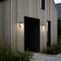 Nordlux Alya udendørs væglampe monteret på en moderne træbeklædt facade ved en døråbning, lyser varmt nedad.