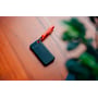 Verbatim Pocket SSD i sort med et tekstureret greb, sat fast i en rød nøglering med et orange bånd, placeret på et træbord med en grøn plante i baggrunden.